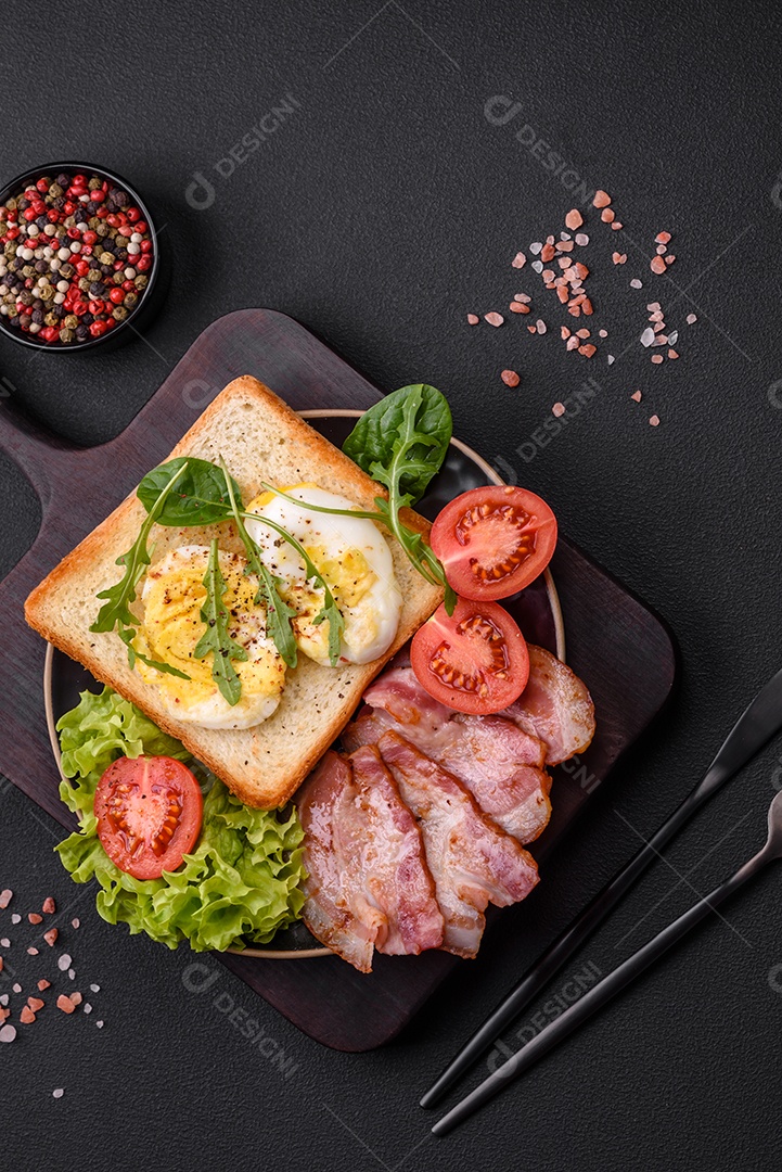 Delicioso almoço saudável composto por bacon, torradas, ovos, tomates, alface e verduras em uma placa preta sobre um fundo de concreto