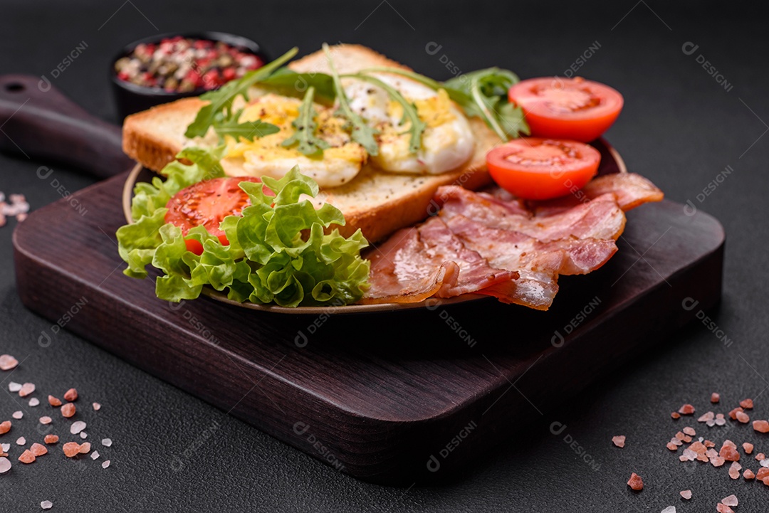 Delicioso almoço saudável composto por bacon, torradas, ovos, tomates, alface e verduras em uma placa preta sobre um fundo de concreto
