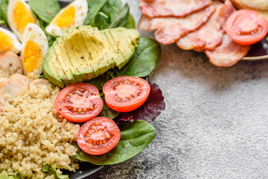 Delicioso almoço saudável composto por frango, abacate, quinoa, ovos, tomate, alface e verduras em uma placa preta sobre fundo de concreto