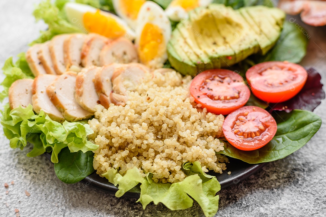 Delicioso almoço saudável composto por frango, abacate, quinoa, ovos, tomate, alface e verduras em uma placa preta sobre fundo de concreto