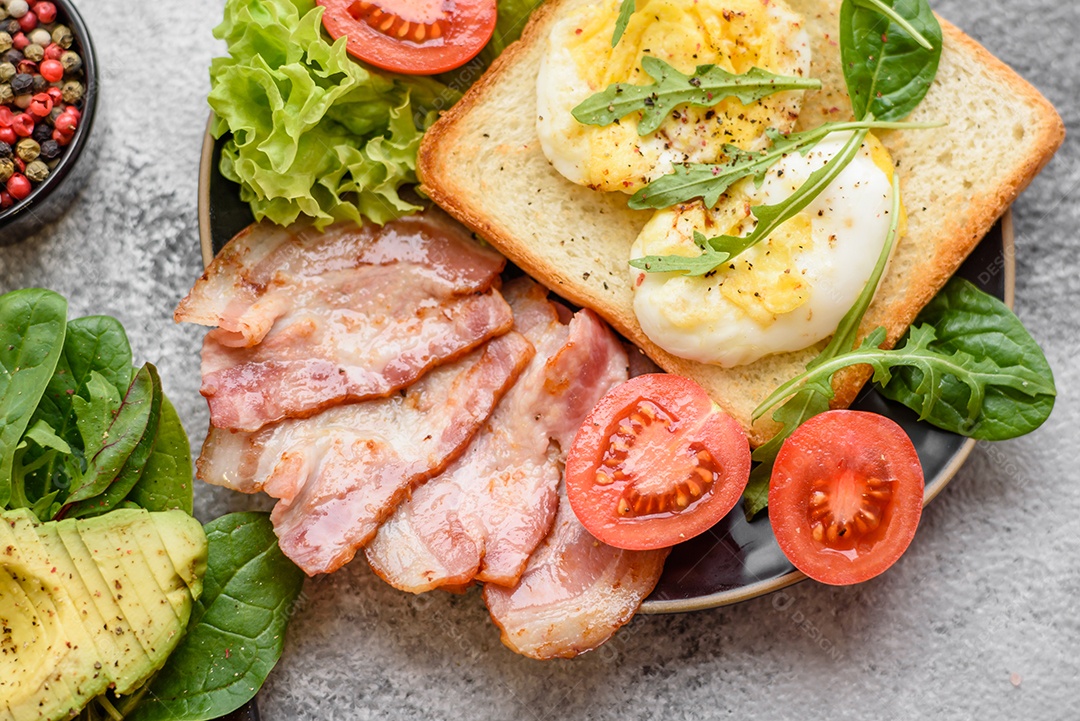 Delicioso almoço saudável composto por bacon, torradas, ovos, tomates, alface e verduras em uma placa preta sobre um fundo de concreto