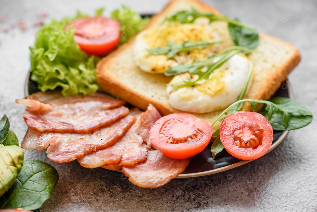 Delicioso almoço saudável composto por bacon, torradas, ovos, tomates, alface e verduras em uma placa preta sobre um fundo de concreto