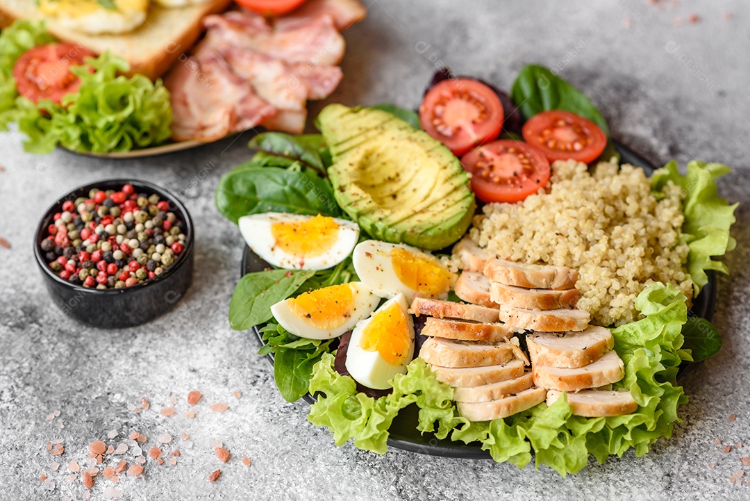 Delicioso almoço saudável composto por frango, abacate, quinoa, ovos, tomate, alface e verduras em uma placa preta sobre fundo de concreto