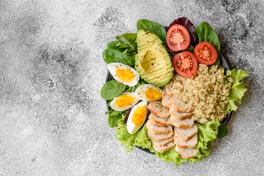 Delicioso almoço saudável composto por frango, abacate, quinoa, ovos, tomate, alface e verduras em uma placa preta sobre fundo de concreto