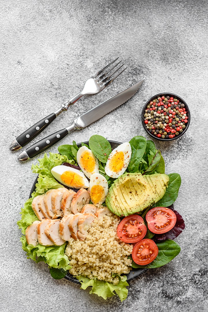 Delicioso almoço saudável composto por frango, abacate, quinoa, ovos, tomate, alface e verduras em uma placa preta sobre fundo de concreto