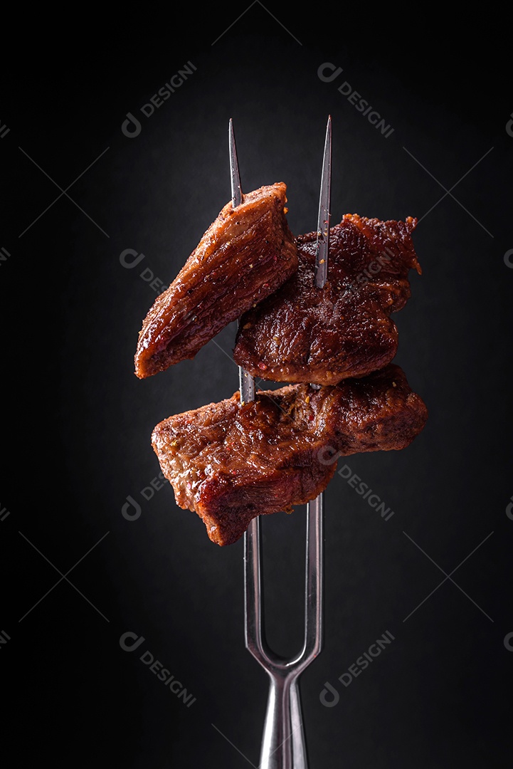 Vários pedaços de carne fresca grelhada com especiarias e ervas e amarrados num garfo sobre um fundo escuro de concreto