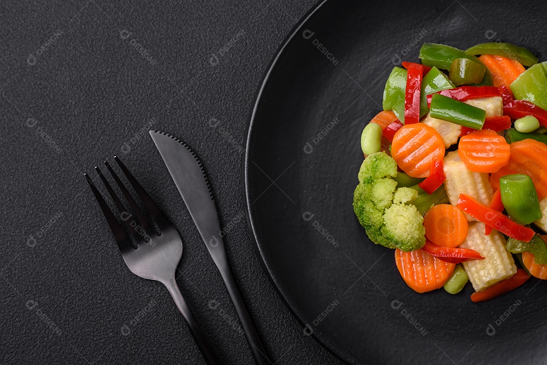 Deliciosos vegetais suculentos de brócolis, cenouras, feijões de espargos, pimentões cozinhados em uma placa preta sobre um fundo escuro de concreto