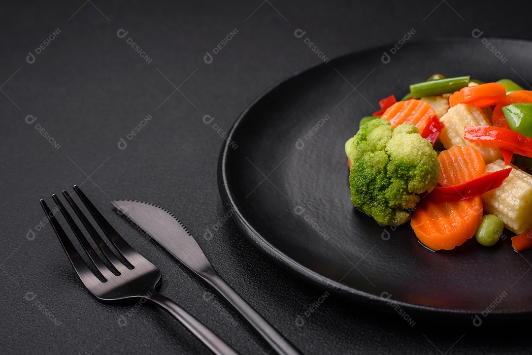 Deliciosos vegetais suculentos de brócolis, cenouras, feijões de espargos, pimentões cozinhados em uma placa preta sobre um fundo escuro de concreto