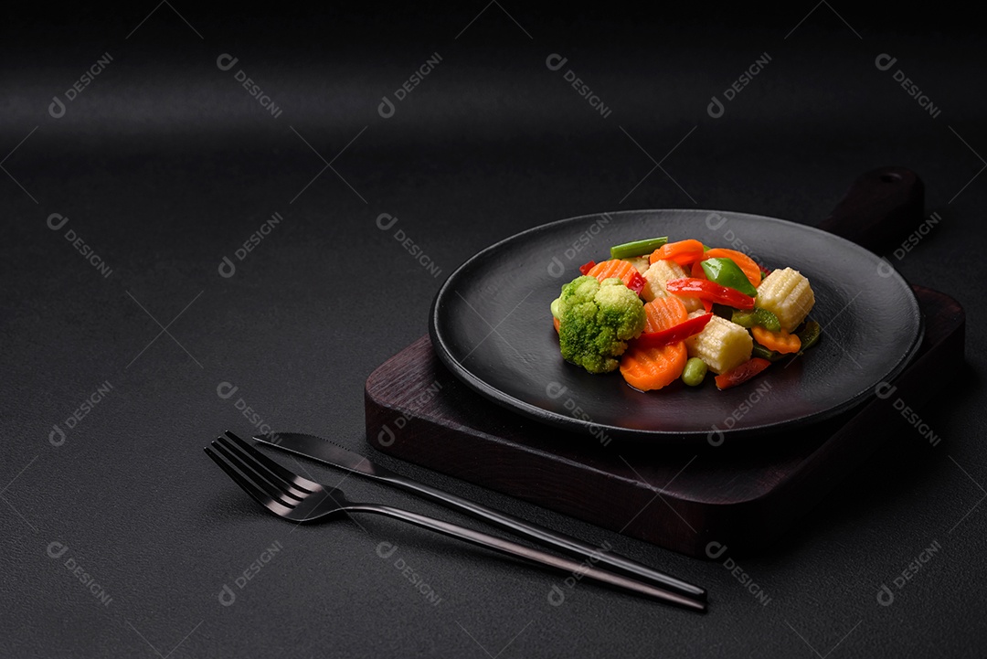 Deliciosos vegetais suculentos de brócolis, cenouras, feijões de espargos, pimentões cozinhados em uma placa preta sobre um fundo escuro de concreto