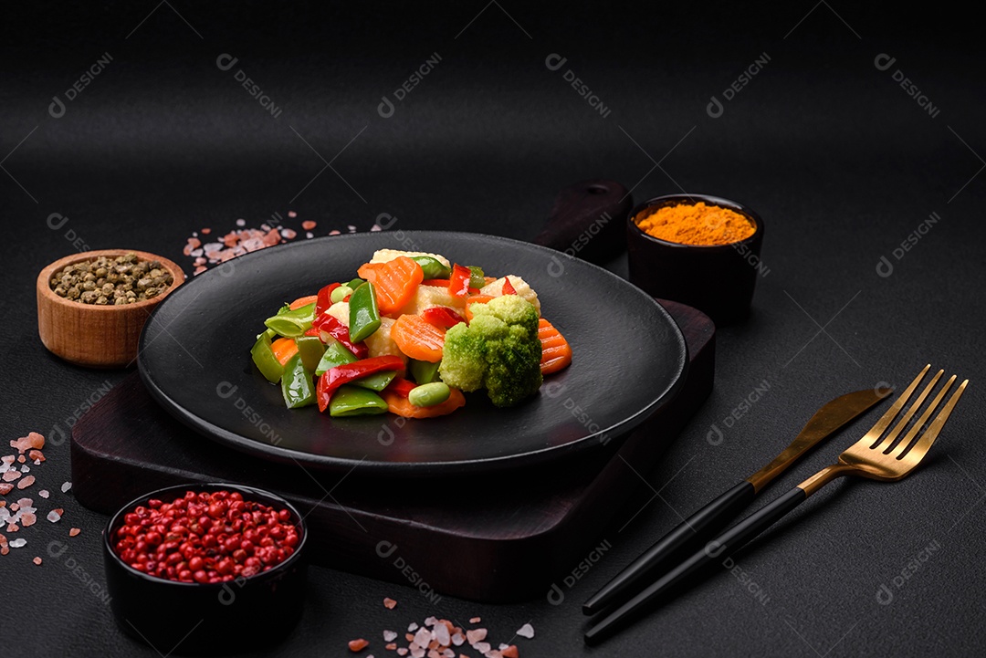 Deliciosos vegetais suculentos de brócolis, cenouras, feijões de espargos, pimentões cozinhados em uma placa preta sobre um fundo escuro de concreto