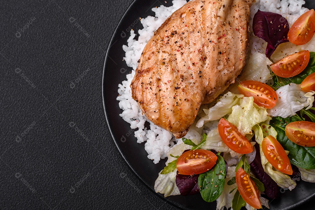 Delicioso café da manhã saudável composto por frango, arroz, verduras e tomate cereja em uma placa de cerâmica preta sobre um fundo escuro de concreto