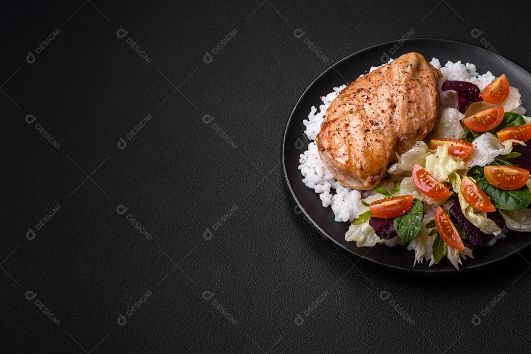 Delicioso café da manhã saudável composto por frango, arroz, verduras e tomate cereja em uma placa de cerâmica preta sobre um fundo escuro de concreto