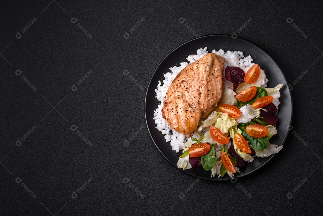 Delicioso café da manhã saudável composto por frango, arroz, verduras e tomate cereja em uma placa de cerâmica preta sobre um fundo escuro de concreto