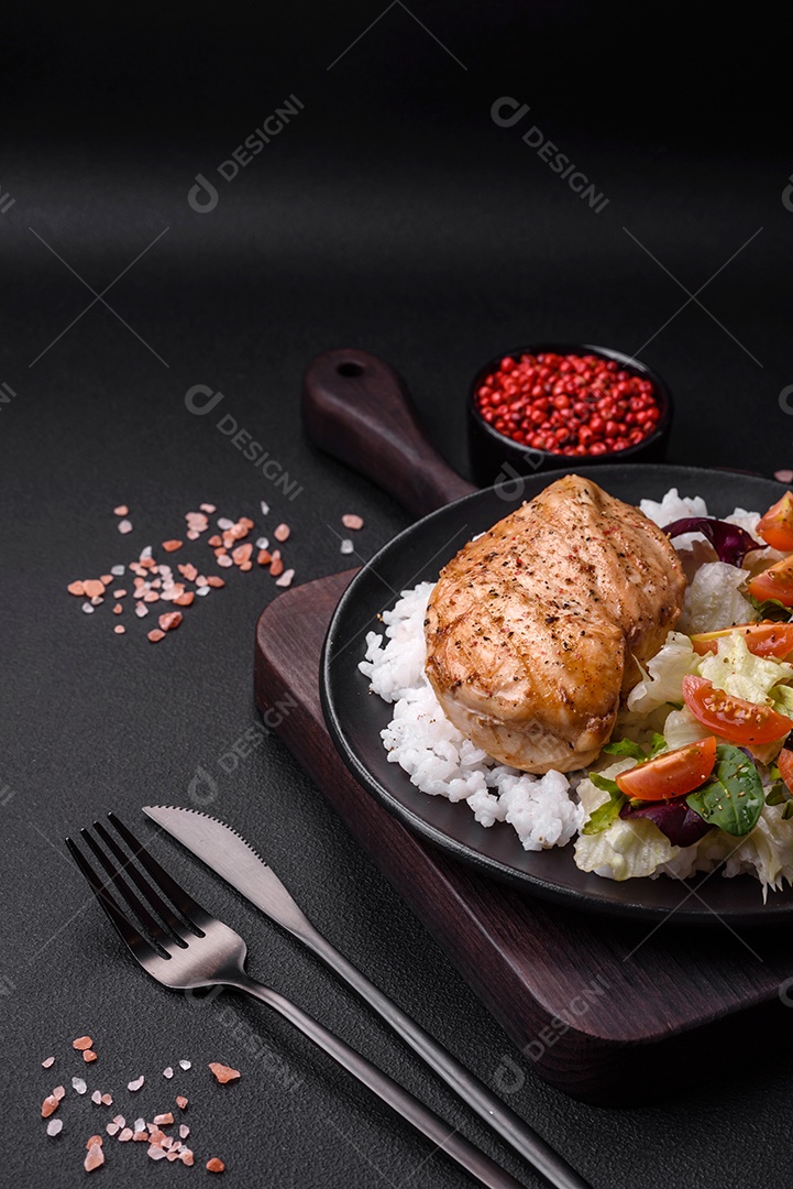 Delicioso café da manhã saudável composto por frango, arroz, verduras e tomate cereja em uma placa de cerâmica preta sobre um fundo escuro de concreto