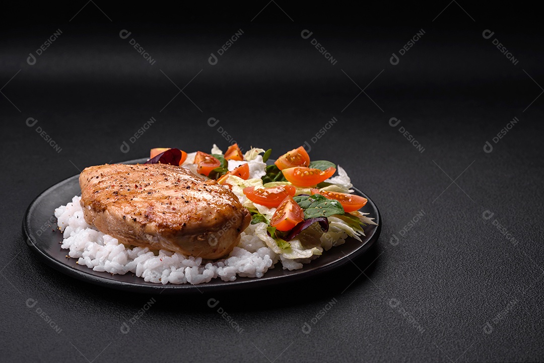 Delicioso café da manhã saudável composto por frango, arroz, verduras e tomate cereja em uma placa de cerâmica preta sobre um fundo escuro de concreto