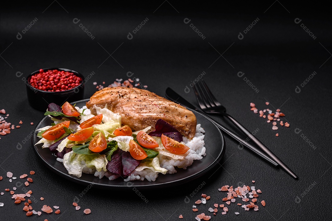 Delicioso café da manhã saudável composto por frango, arroz, verduras e tomate cereja em uma placa de cerâmica preta sobre um fundo escuro de concreto