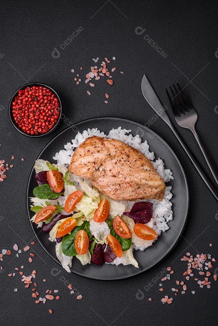 Delicioso café da manhã saudável composto por frango, arroz, verduras e tomate cereja em uma placa de cerâmica preta sobre um fundo escuro de concreto
