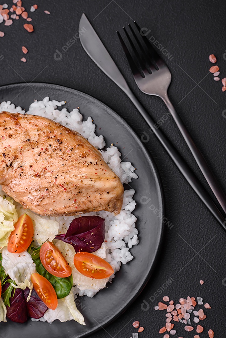 Delicioso café da manhã saudável composto por frango, arroz, verduras e tomate cereja em uma placa de cerâmica preta sobre um fundo escuro de concreto