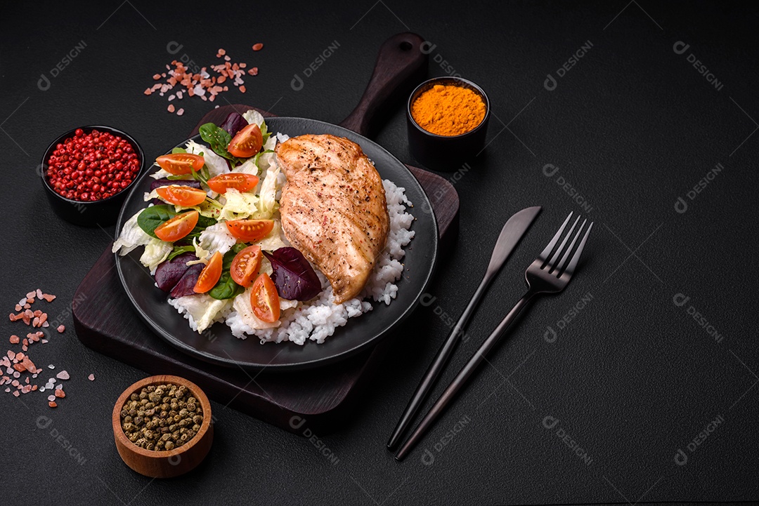 Delicioso café da manhã saudável composto por frango, arroz, verduras e tomate cereja em uma placa de cerâmica preta sobre um fundo escuro de concreto