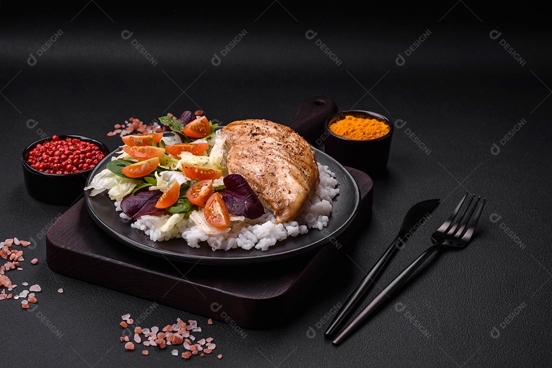 Delicioso café da manhã saudável composto por frango, arroz, verduras e tomate cereja em uma placa de cerâmica preta sobre um fundo escuro de concreto