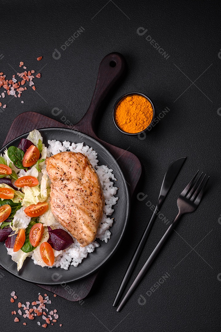 Delicioso café da manhã saudável composto por frango, arroz, verduras e tomate cereja em uma placa de cerâmica preta sobre um fundo escuro de concreto