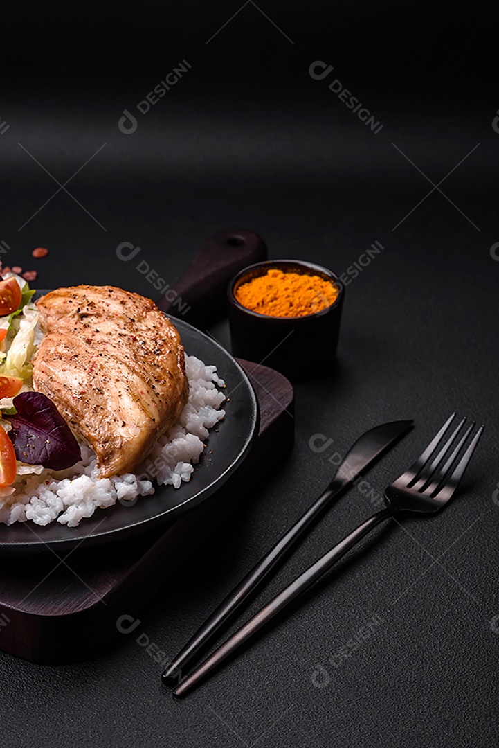 Delicioso café da manhã saudável composto por frango, arroz, verduras e tomate cereja em uma placa de cerâmica preta sobre um fundo escuro de concreto