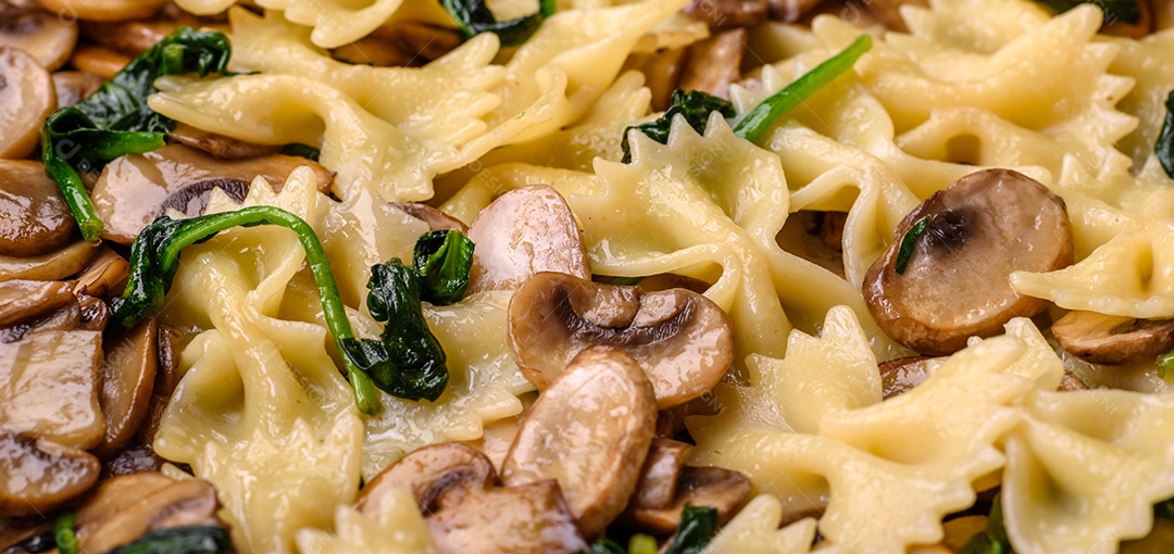 Deliciosa massa farfalle com cogumelos, queijo e espinafre com especiarias em uma placa preta sobre um fundo escuro de concreto