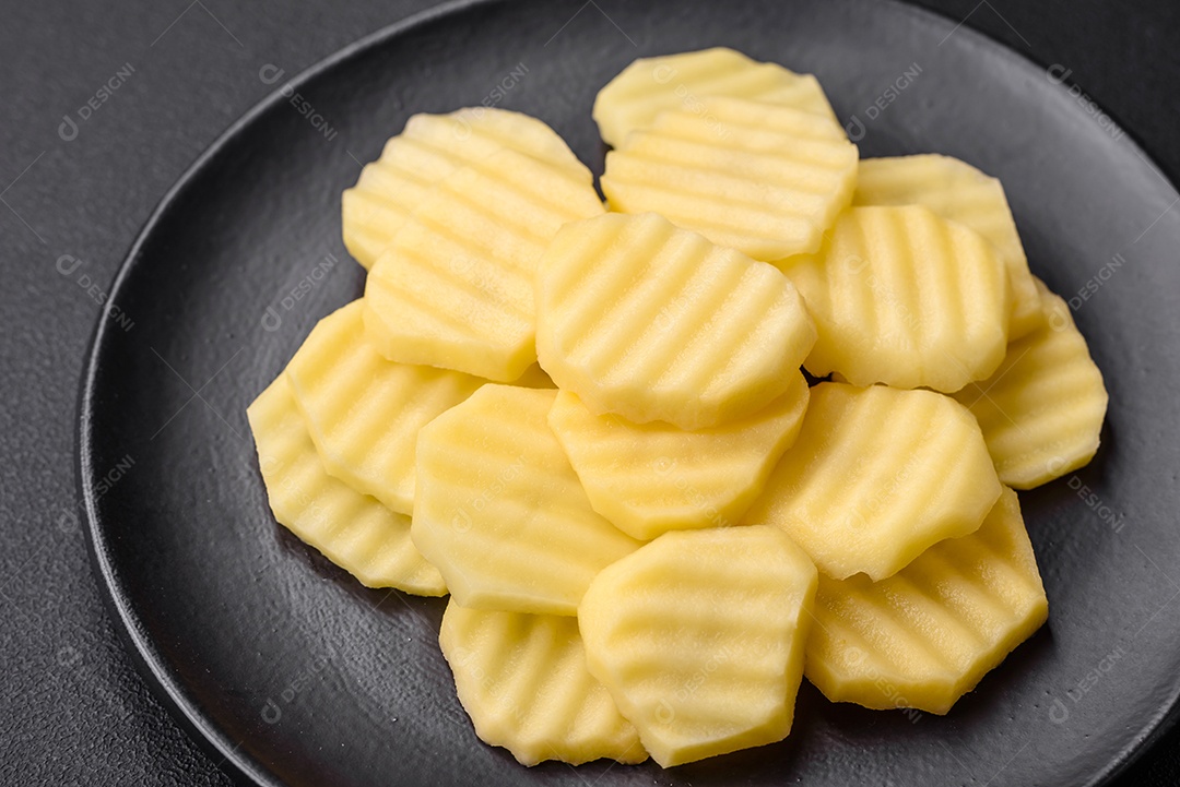 Batatas frescas cruas cortadas em fatias numa placa de cerâmica preta sobre um fundo escuro de concreto