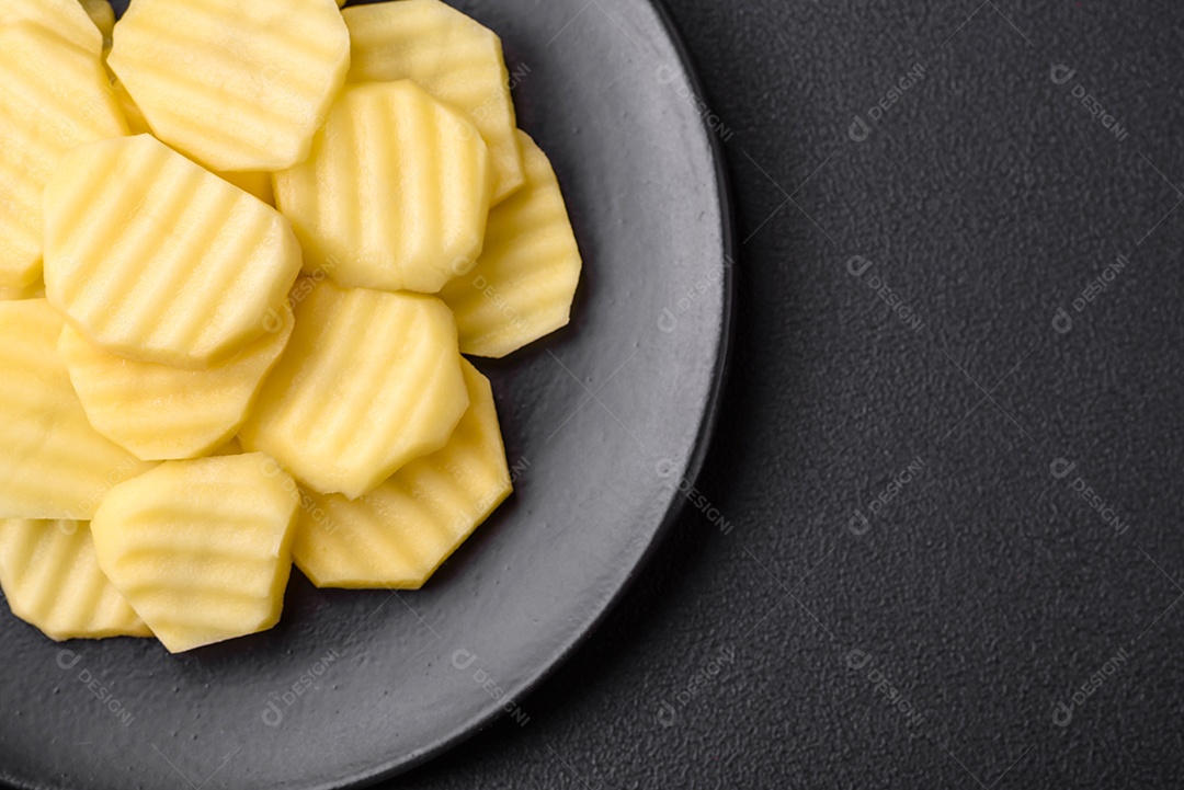 Batatas frescas cruas cortadas em fatias numa placa de cerâmica preta sobre um fundo escuro de concreto