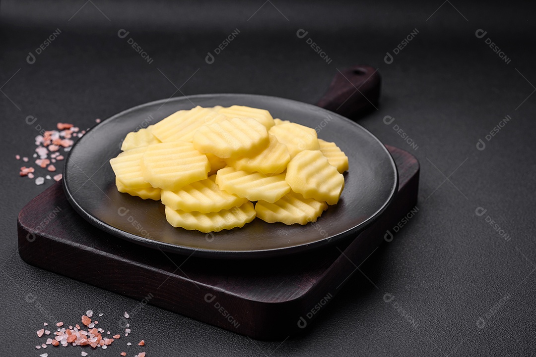 Batatas frescas cruas cortadas em fatias numa placa de cerâmica preta sobre um fundo escuro de concreto