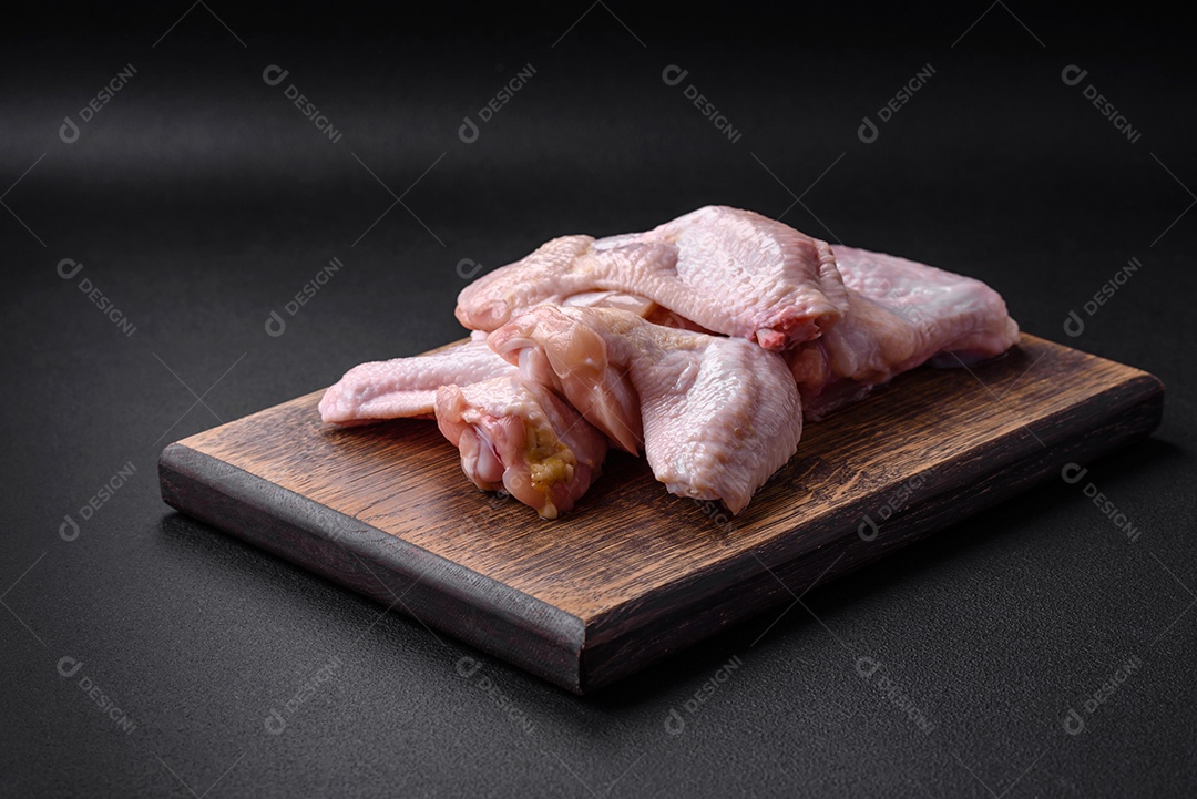 Asas de frango cru com sal, especiarias e ervas em uma tábua de madeira sobre um fundo escuro de concreto