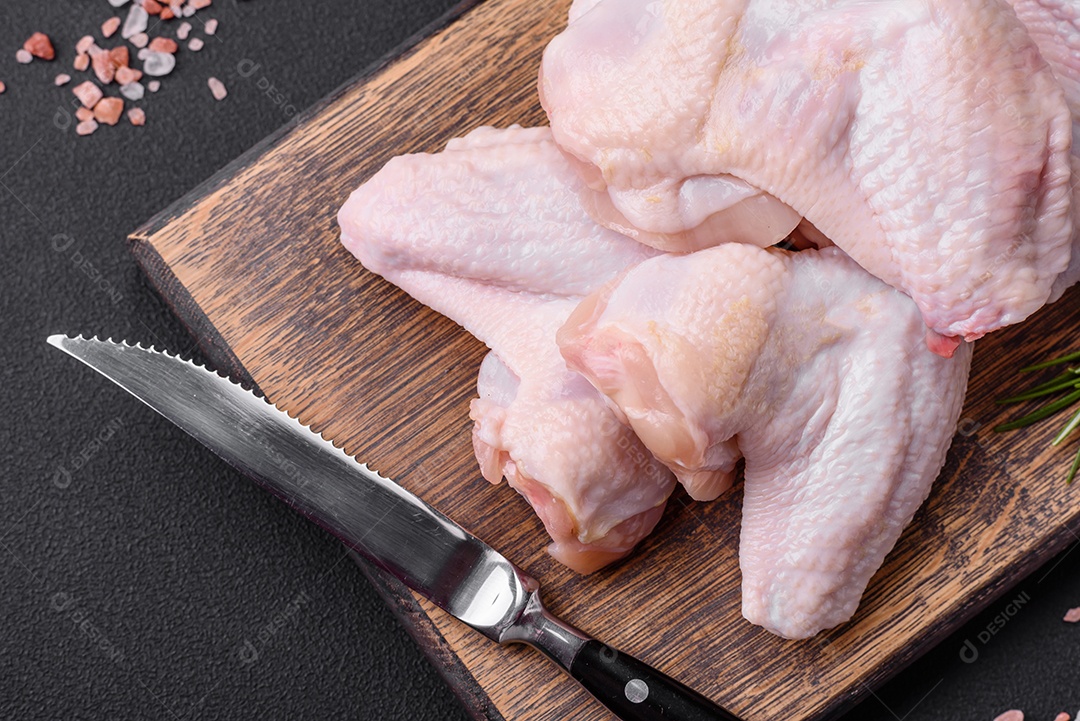Asas de frango cru com sal, especiarias e ervas em uma tábua de madeira sobre um fundo escuro de concreto