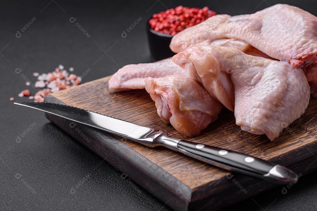 Asas de frango cru com sal, especiarias e ervas em uma tábua de madeira sobre um fundo escuro de concreto