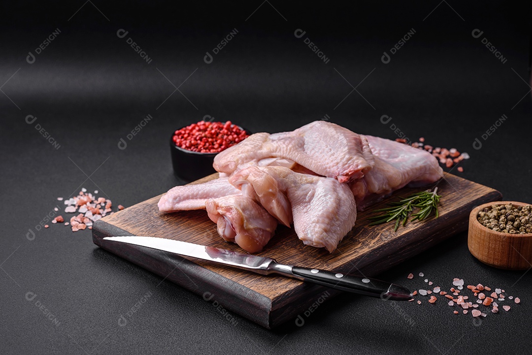 Asas de frango cru com sal, especiarias e ervas em uma tábua de madeira sobre um fundo escuro de concreto
