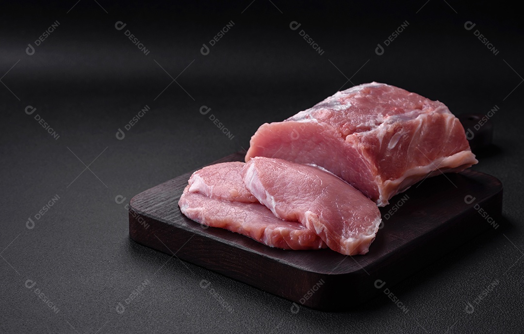 Carne de porco fresca e suculenta sobre uma tábua de madeira com especiarias e sal sobre um fundo escuro de concreto