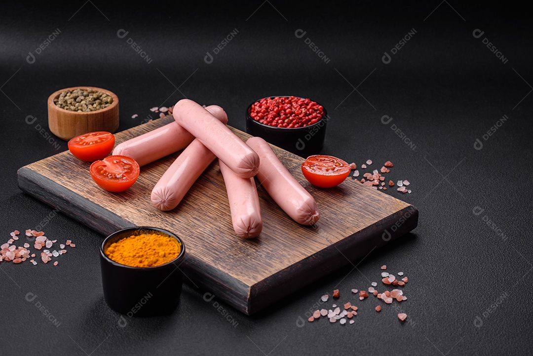 Salsichas cozidas em uma tábua de madeira com tomate, especiarias e sal sobre um fundo escuro de concreto