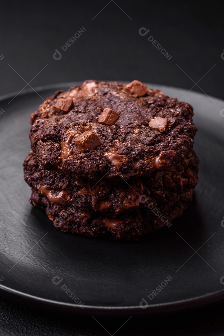 Deliciosos biscoitos redondos de aveia fresca com chocolate em uma placa de cerâmica preta sobre um fundo escuro de concreto