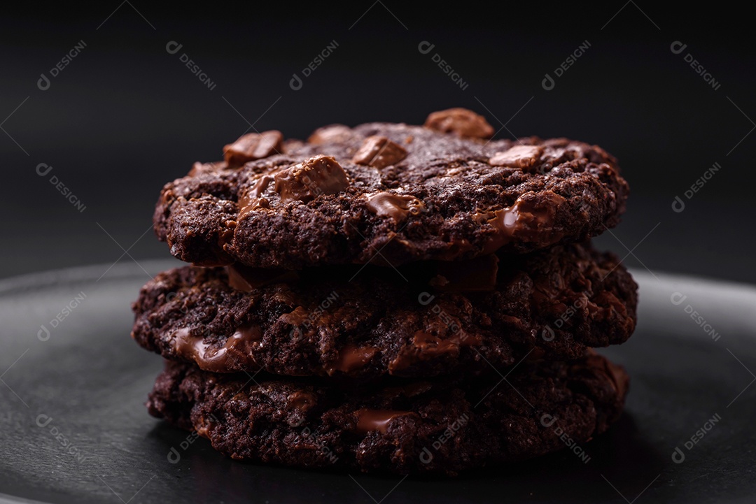 Deliciosos biscoitos redondos de aveia fresca com chocolate em uma placa de cerâmica preta sobre um fundo escuro de concreto
