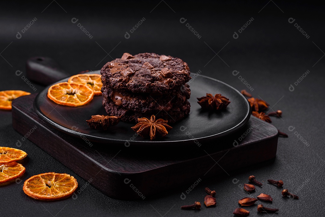 Deliciosos biscoitos redondos de aveia fresca com chocolate em uma placa de cerâmica preta sobre um fundo escuro de concreto