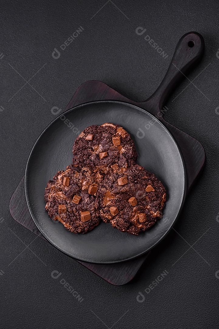 Deliciosos biscoitos redondos de aveia fresca com chocolate em uma placa de cerâmica preta sobre um fundo escuro de concreto