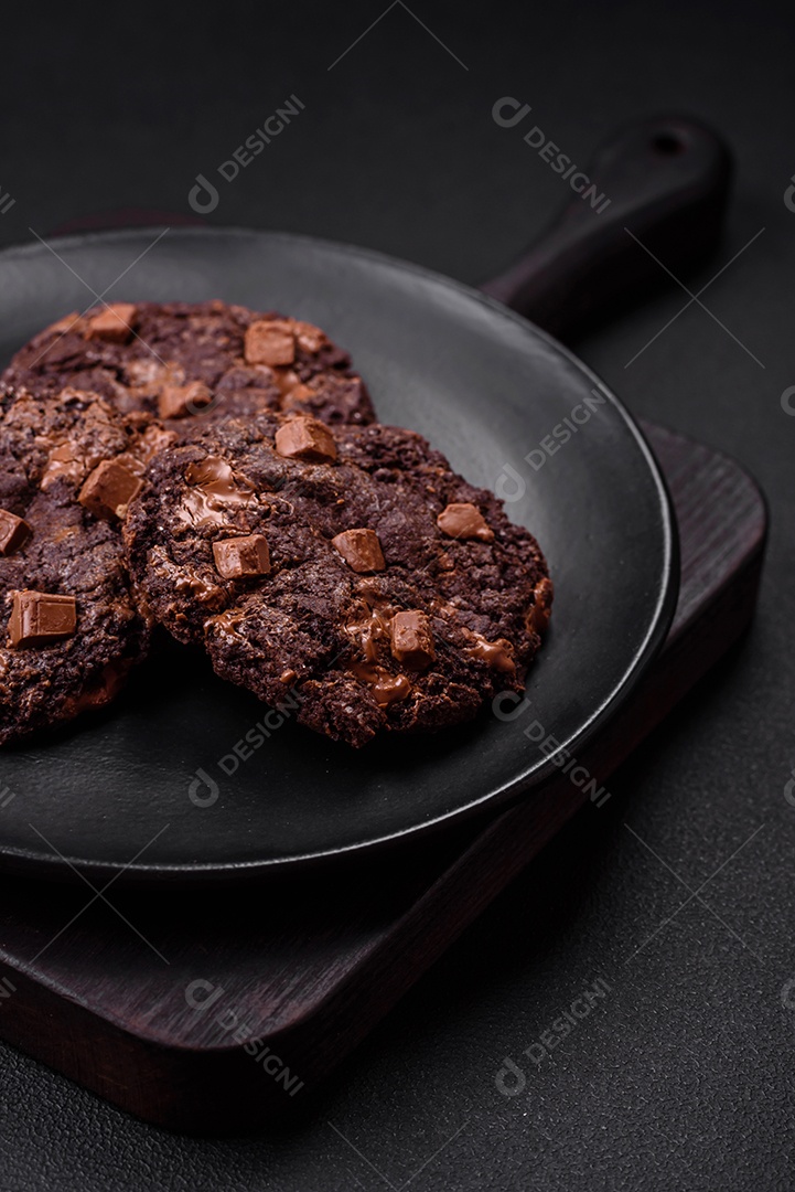 Deliciosos biscoitos redondos de aveia fresca com chocolate em uma placa de cerâmica preta sobre um fundo escuro de concreto