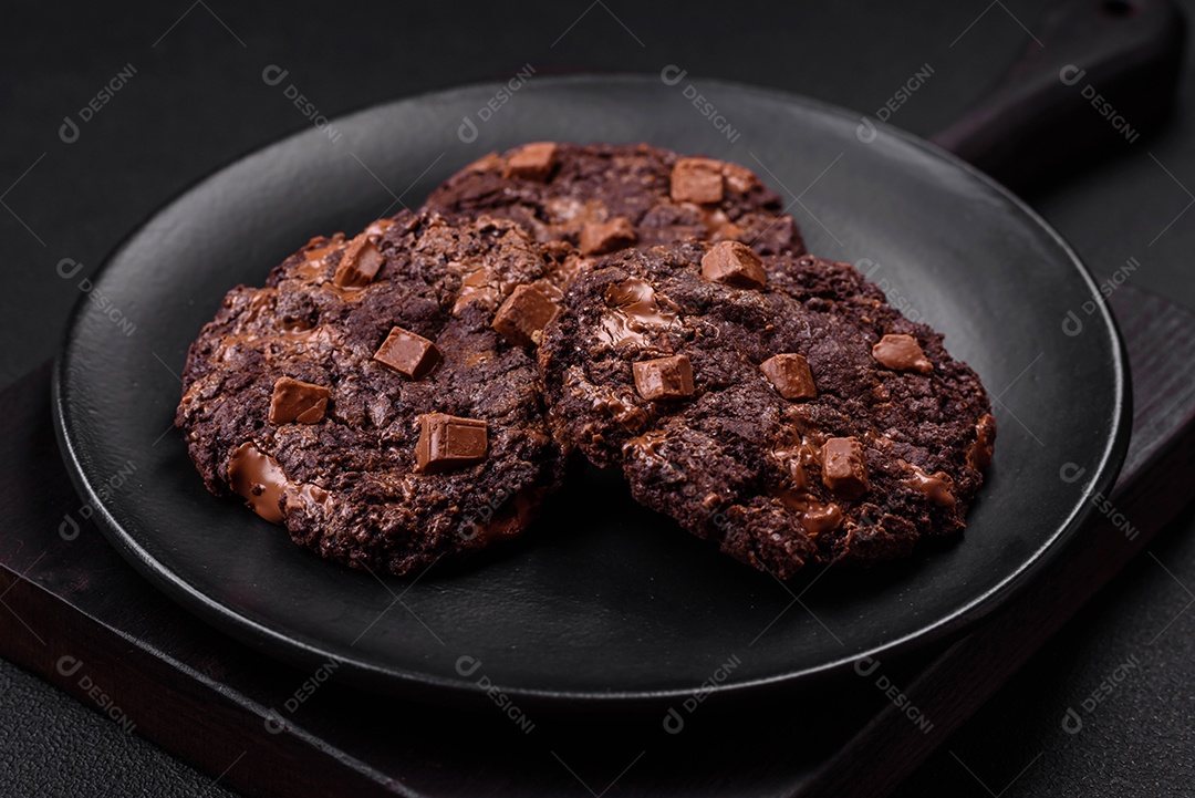 Deliciosos biscoitos redondos de aveia fresca com chocolate em uma placa de cerâmica preta sobre um fundo escuro de concreto