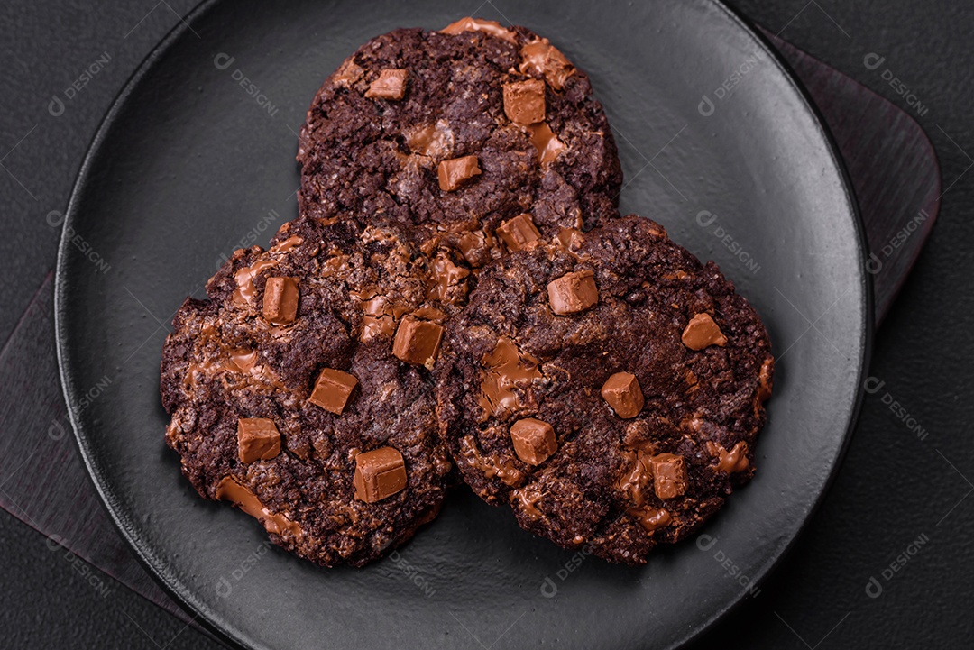 Deliciosos biscoitos redondos de aveia fresca com chocolate em uma placa de cerâmica preta sobre um fundo escuro de concreto