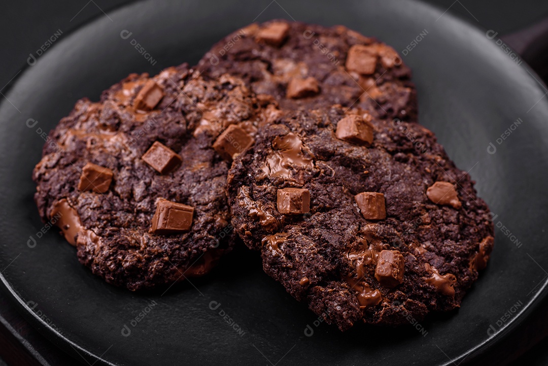 Deliciosos biscoitos redondos de aveia fresca com chocolate em uma placa de cerâmica preta sobre um fundo escuro de concreto