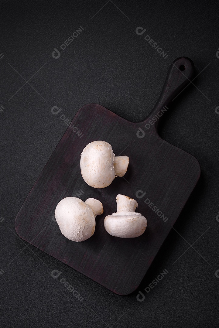 Cogumelos champignon crus frescos sobre uma tábua de madeira com especiarias e ervas sobre um fundo escuro de concreto