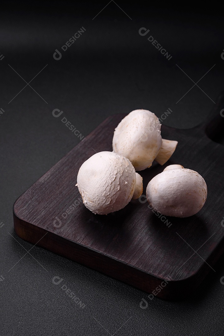 Cogumelos champignon crus frescos sobre uma tábua de madeira com especiarias e ervas sobre um fundo escuro de concreto