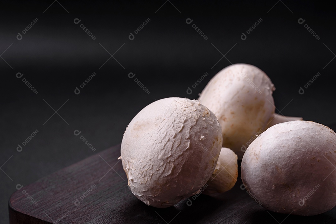 Cogumelos champignon crus frescos sobre uma tábua de madeira com especiarias e ervas sobre um fundo escuro de concreto
