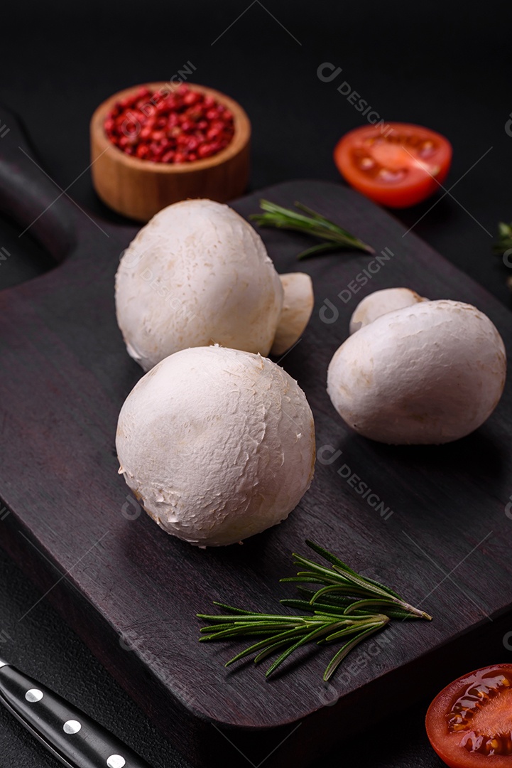 Cogumelos champignon crus frescos sobre uma tábua de madeira com especiarias e ervas sobre um fundo escuro de concreto