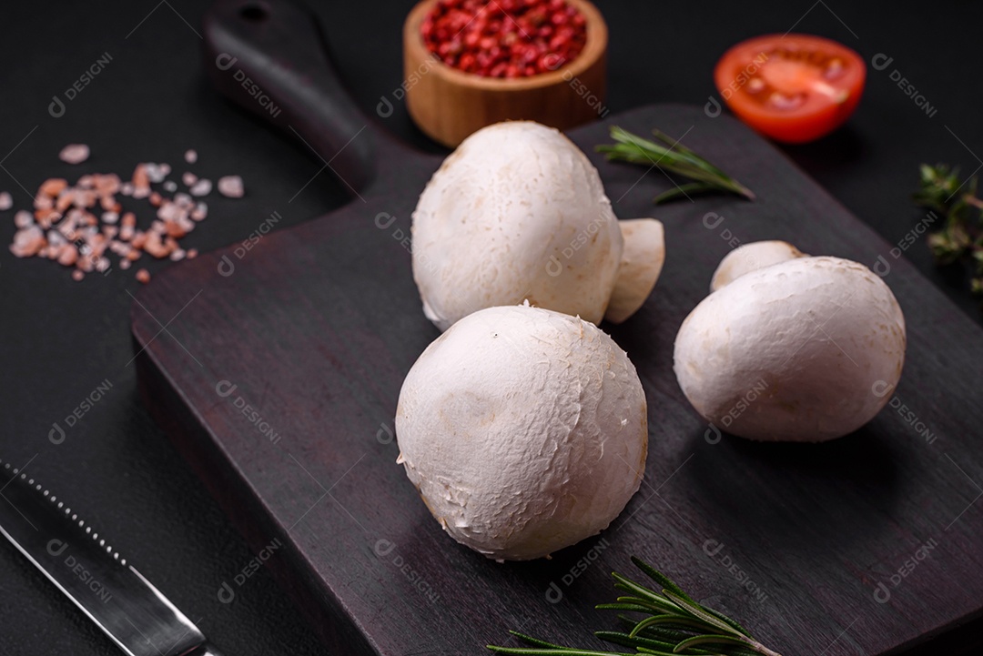 Cogumelos champignon crus frescos sobre uma tábua de madeira com especiarias e ervas sobre um fundo escuro de concreto