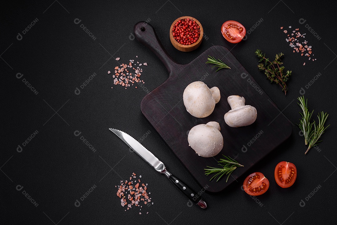 Cogumelos champignon crus frescos sobre uma tábua de madeira com especiarias e ervas sobre um fundo escuro de concreto